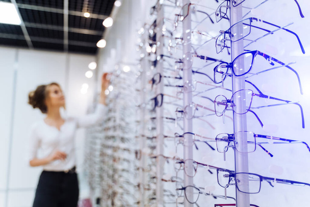 Eyeglasses shop. Stand with glasses in the store of optics. Woman on blurred background chooses glasses. - Foto, Imagen
