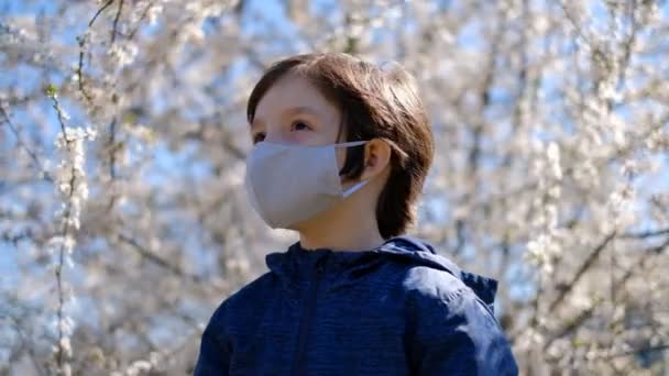 小さな男の子が家を出て、隔離中の保護マスクとコロナウイルスの流行の間に散歩しました。保護マスクの開花木の近くの公園に男の子が立っている. - 映像、動画