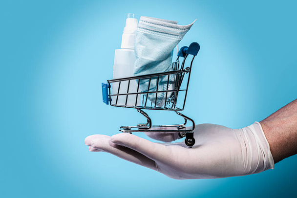 Shopping cart with surgical mask and hand sanitizer fluid hold up by and human hand wearing gloves on a light blue background. - Photo, Image