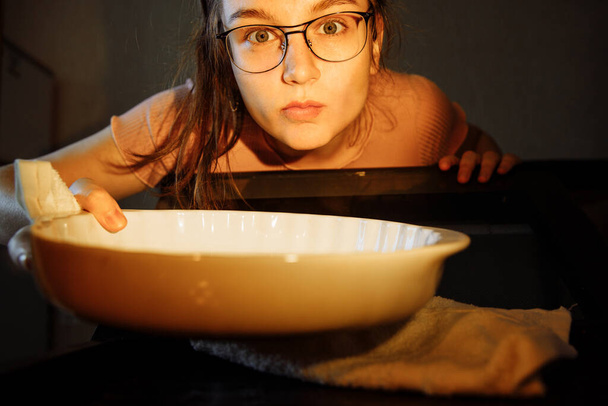 View from oven. The girl takes a glass pan out of the oven. The person is illuminated by the light from the oven lamp. - Fotó, kép