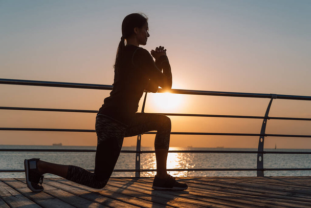 Young athletic girl performs squats on wooden embankment by the sea in early morning. Healthy lifestyle, coaching, training concept. - Фото, зображення