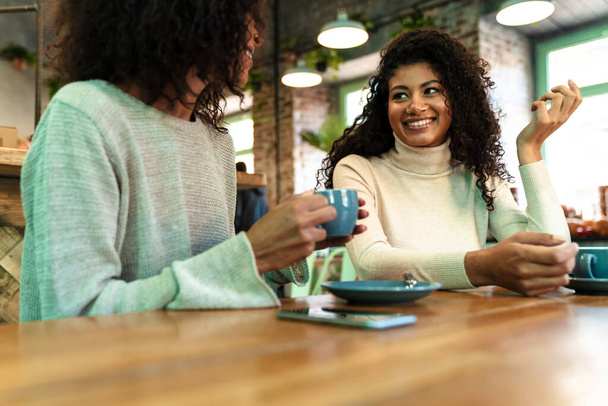 Two cheerful young african girlfriends sitting at the cafe drinking coffee, talking - 写真・画像