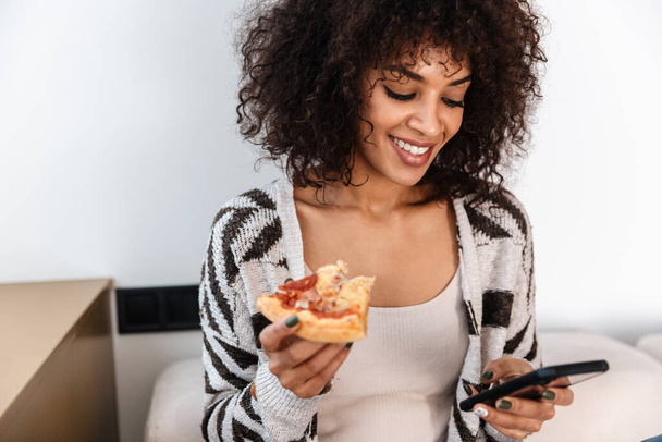 Image of happy positive pretty young african woman indoors at home eat pizza using mobile phone. - 写真・画像