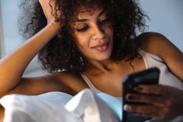 Image of positive optimistic young african woman indoors at home using mobile phone lies in bed. - Photo, Image
