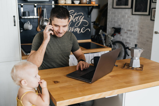 Attractive man in the interior of a modern rustic kitchen working on a laptop with his daughter child. remote work. - Zdjęcie, obraz
