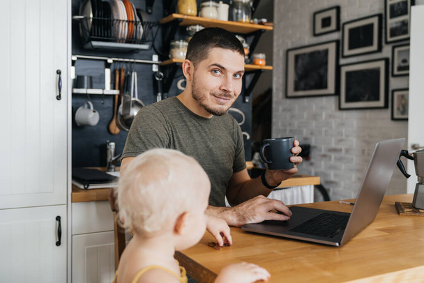 Thuis werken op afstand. Positieve Portret van een man die werkt op een laptop in de keuken interieur met zijn dochter. - Foto, afbeelding