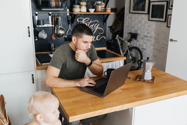 Trabajo remoto en casa. Hombre padre trabajando en un ordenador portátil en el interior de la cocina con su hija
. - Foto, Imagen