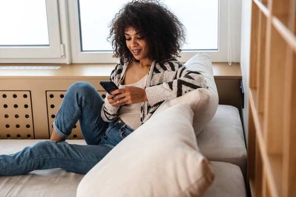 Image of positive optimistic young african woman indoors at home using mobile phone. - Фото, изображение