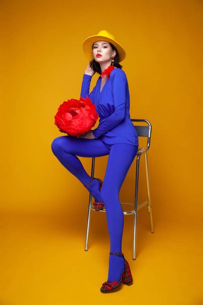 Lebendiges Modeporträt. junges schönes Model posiert auf gelbem Hintergrund. helles Konzept. Frau in blauem Kostüm und gelbem Hut. Kontrastprogramm kreatives Foto. Dame sitzt auf Stuhl - Foto, Bild
