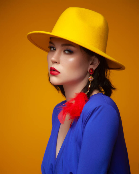 fiatal gyönyörű nő pózol a stúdióban. Fényes divatkoncepció. lány göndör barna haj visel kék ruhát pózol sárga háttér. Sárga kalap. modell piros ajkakkal és kék szemceruzával - Fotó, kép