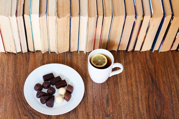 cup of tea and chocolate candies on white plate, stack of old book on wooden table, education concept background, many books piles with copy space for text - Foto, afbeelding
