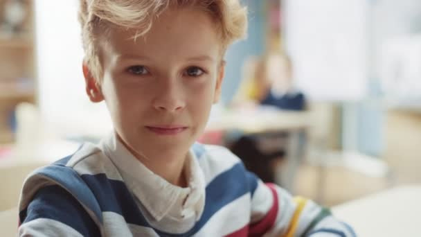 Aranyos kisfiú, hullámos szőke hajjal, aki az iskolaasztalánál ül, boldogan vigyorog. Okos kisfiú bájos mosollyal ül az osztályteremben. Közelkép Kamera Shot - Felvétel, videó