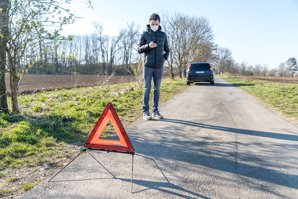 triángulo rojo de advertencia en la carretera frente a un coche roto. Un joven usando un teléfono para pedir servicio de asistencia
 - Foto, imagen