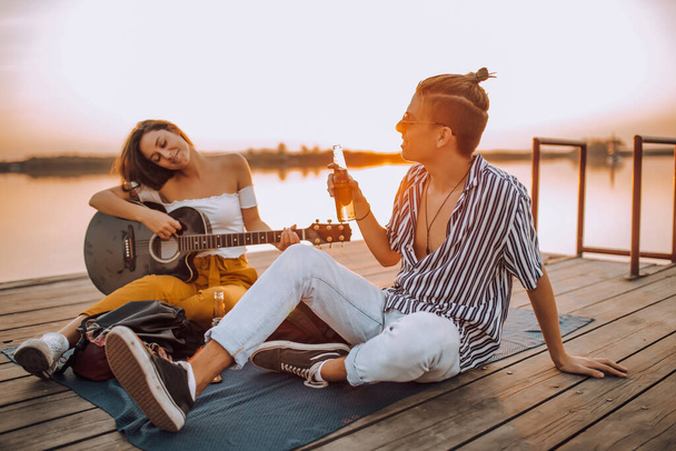 Mutlu genç çift bira içiyor, gitar çalıyor ve nehir kenarında şarkı söylüyor. - Fotoğraf, Görsel