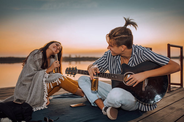 Fröhliches junges Paar trinkt Bier, spielt Gitarre und singt, während es auf einem hölzernen Ponton in der Nähe eines Flusses sitzt. - Foto, Bild