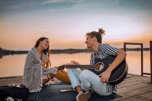 Happy cheerful young couple drinking beer, playing a guitar and singing while sitting on a wooden pontoon near a river. - Photo, Image