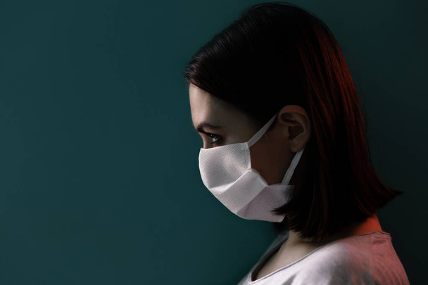 isolated girl in medical mask close-up in profile, dramatic portrait, against the wall, red background light - Foto, afbeelding