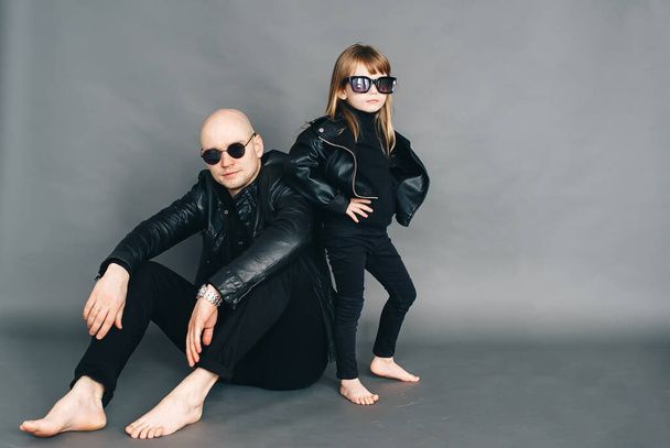 Handsome bald father with his little daughter in black sunglasses in biker clothes are posing in studio. Horizontal portrait - Fotoğraf, Görsel