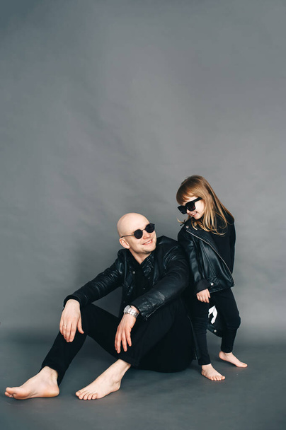 Handsome bald father with his little daughter in black sunglasses in biker clothes are posing in studio. Vertical portrait - Photo, Image