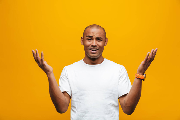 Portrait of an attractive confused casual young african man standing over yellow background, gesturing - Foto, Bild