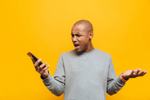 Портрет привлекательного сердитого молодого африканца, стоящего на желтом фоне, держащего в руках мобильный телефон
 - Фото, изображение