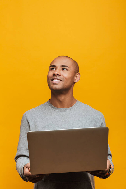 Portrait of an attractive smiling confident casual young african man standing over yellow background, holding laptop computer, looking away - Zdjęcie, obraz