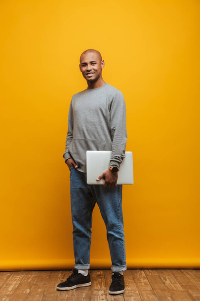 Full length portrait of an attractive happy confident casual young man standing over yellow background, carrying laptop computer - Фото, изображение
