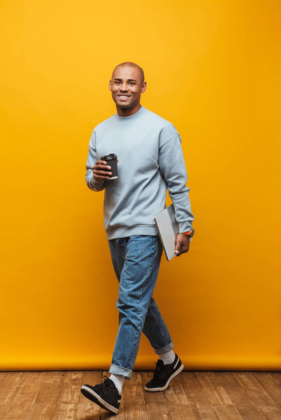 Full length portrait of an attractive happy confident casual young man walking over yellow background, carrying laptop computer, drinking takeaway coffee - Foto, imagen