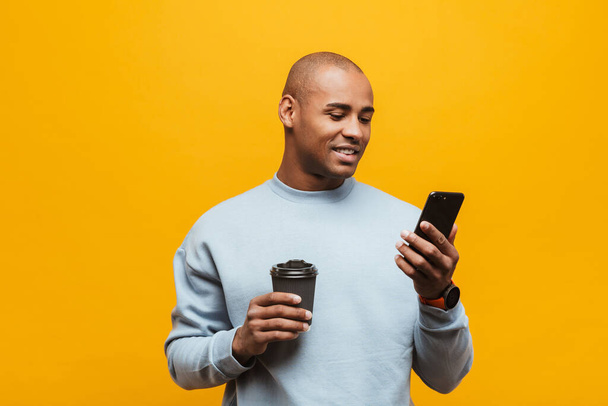 Portrait of an attractive smiling confident casual young african man standing over yellow background, using mobile phone - Foto, afbeelding
