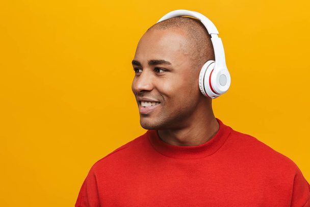 Portrait of an attractive smiling confident casual young african man standing over yellow background, listening to music with wireless headphones, looking away - Photo, Image
