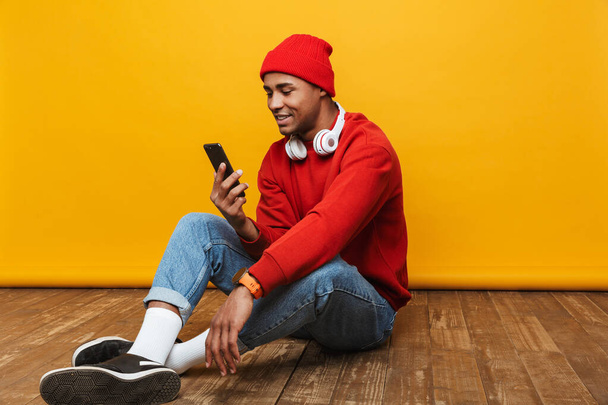 Portrait of an attractive happy casual young african man sitting on a floor over yellow background - Φωτογραφία, εικόνα
