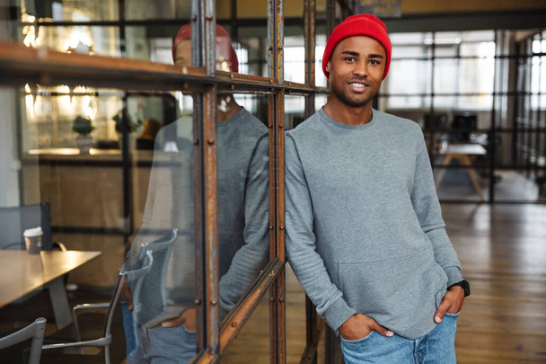 Image of young attractive african american man wearing hat standing with arms at pockets while working in office - Foto, Imagem