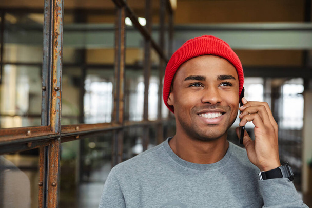 Image of young attractive african american man wearing hat talking on cellphone while working in office - Photo, Image