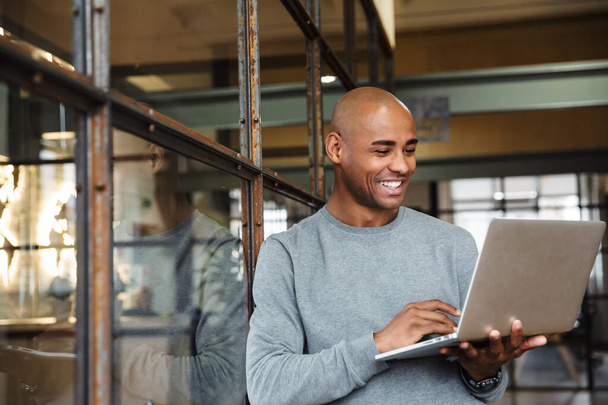 Imagem do jovem homem americano africano careca atraente vestindo camisola segurando computador portátil enquanto trabalhava no escritório
 - Foto, Imagem