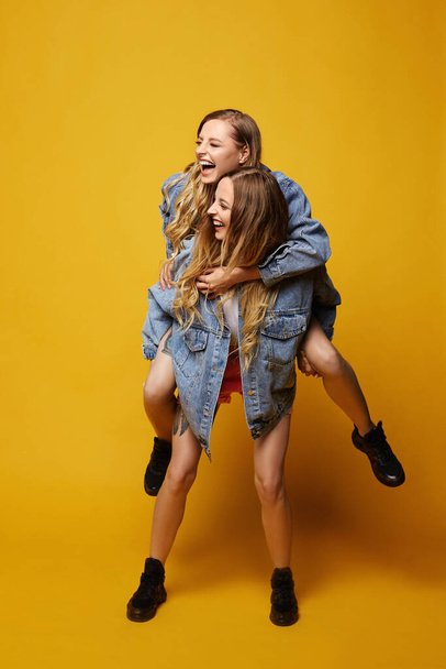 Young women girlfriends in summer outfits have fun and posing at the yellow background. Summertime lifestyle. Full-length portrait of two female models, summer fashion concept - Photo, Image