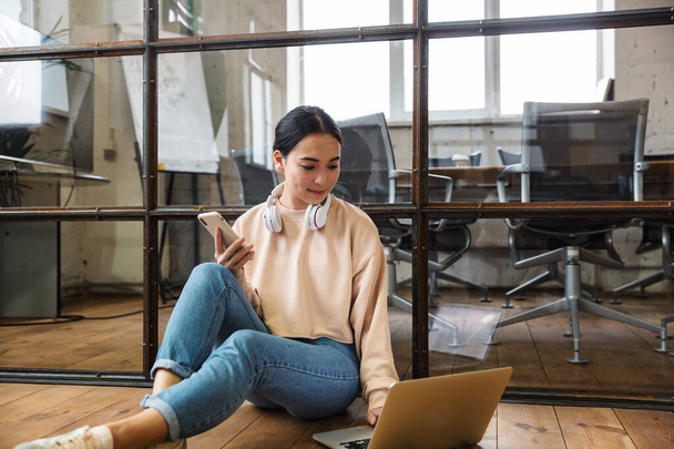 Image of a positive asian young woman sit indoors on floor using laptop computer and mobile phone. - Foto, Bild