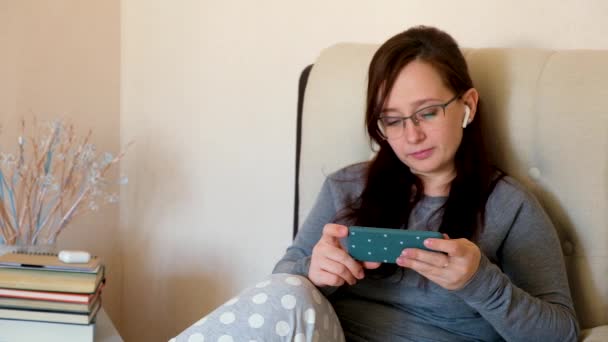 Usmívající se uvolněná žena drží smartphone a dívá se na video, jak sedí v obývacím pokoji na posteli. Šťastná dívka s brýlemi a sluchátky při pohledu na mobilní telefon. - Záběry, video