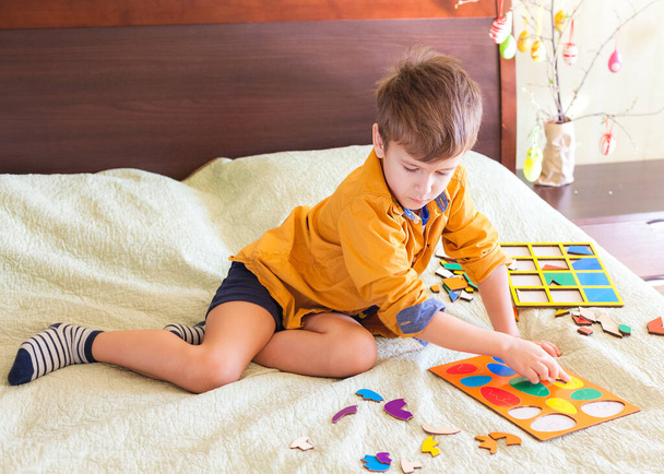 Boy collects wooden puzzle in the form of Easter eggs, home interior. Easter decoration in the background - Foto, Bild