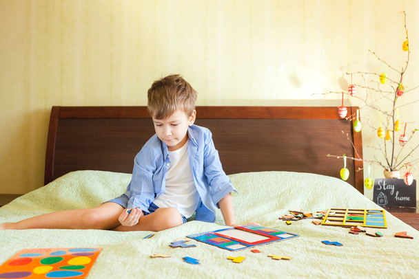 Boy collects wooden puzzle sitting on bed at home interior. Easter decoration at background and text Stay home. Being home concept at Easter holiday - Foto, imagen