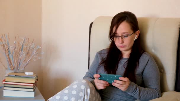 Blanke vrolijke jonge vrouw rusten in de woonkamer met smartphone in handen. Vrouwelijke tikken, scrollen en surfen online. Dame kijkt naar iets aan de telefoon. - Video