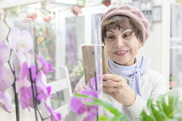 Happy caucasian elderly woman in a beret, smiling, photographs orchid flowers on the phone - Photo, Image