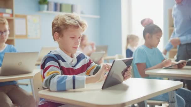 Informatikunterricht in der Grundschule: Netter Junge nutzt digitalen Tablet-Computer, seine Klassenkameraden arbeiten auch mit Laptops. Kinder erhalten moderne Bildung in MINT-Fächern, Spielen und Lernen - Filmmaterial, Video