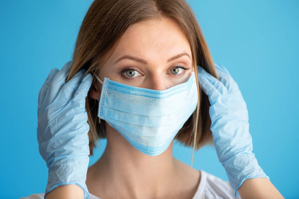 Νοσοκόμα ή γιατρός με μάσκα προσώπου και γάντια. Ιατρική περίθαλψη, χειρουργική επέμβαση. Κοντινό πορτραίτο νεαρής καυκάσιας γυναίκας σε μπλε φόντο - Φωτογραφία, εικόνα