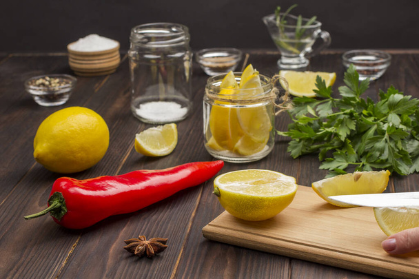 Sliced lemon on cutting board. Hands chop a lemon on cutting board. Lemon in jar. Chili pepper, whole lemon and salt on table. Natural source of strengthening immunity. Dark Wood background - Fotoğraf, Görsel