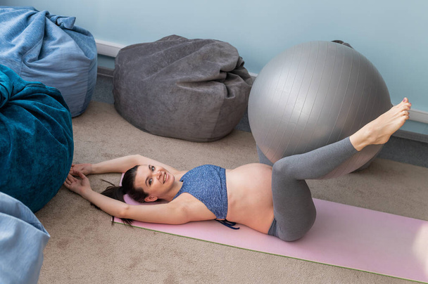 Pregnant european woman performs exercises with fitness ball. Expectant mother is doing yoga in the 3rd trimester. Girl in the classroom in preparation for childbirth. - Foto, Imagen