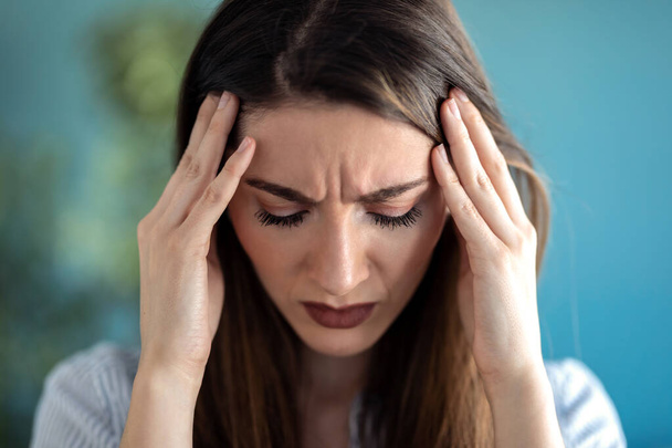 Portret van gefrustreerde jonge vrouw met hoofdpijn en fronsen terwijl ze thuis staat. - Foto, afbeelding