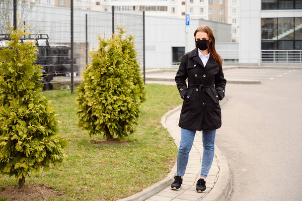 Porträt eines kaukasischen brünetten Mädchens auf einer Straße in der Stadt mit einer medizinischen schwarzen Maske, während sie draußen in einem schwarzen Mantel spaziert - Foto, Bild