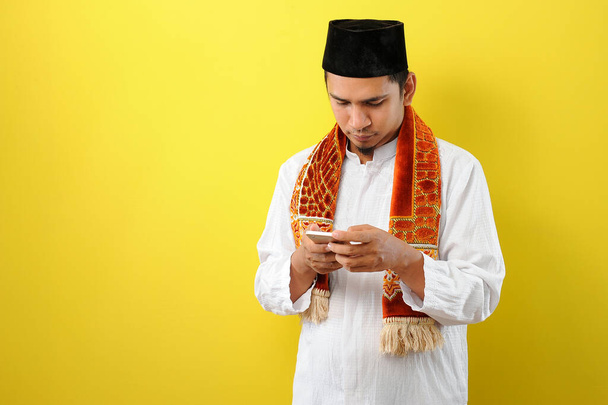 Jeune homme musulman asiatique portant des vêtements moslim tenant téléphone portable regarder l'écran de téléphone, isolé sur fond jaune
 - Photo, image