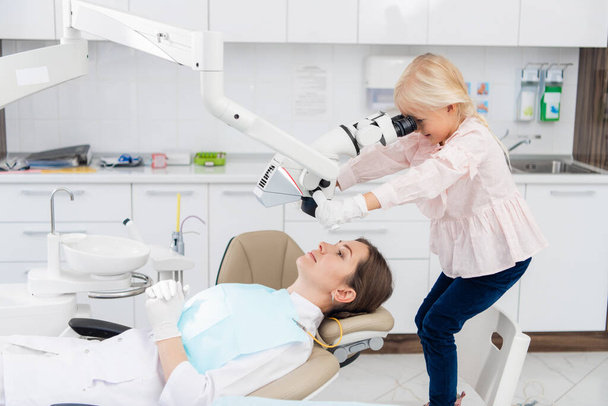 Urocza dziewczynka wykonująca badanie lekarskie kobiety, lekarz leży na krześle dentystycznym. Dentysta i dziecko w gabinecie dentystycznym. Mała dziewczynka bawiąca się w gabinecie dentystycznym - Zdjęcie, obraz
