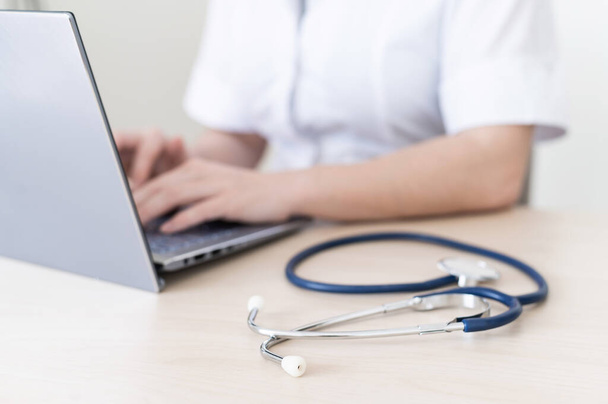 Médica feminina irreconhecível em uniforme usa um laptop. Um médico sem rosto escreve um relatório sobre o computador na secretária. Cultivadas
. - Foto, Imagem
